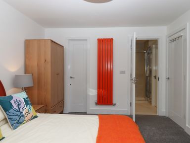 A bedroom with a bed with colorful pillows and an orange blanket a wooden wardrobe a bedside table with a lamp a bright orange wall radiator and a door leading to a bathroom at Rolles Rainbow in Exmouth