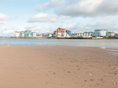 A sandy beach with scattered small rocks and water with colorful buildings on the opposite shore at Rolles Rainbow in Exmouth