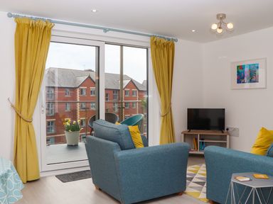 A living room with blue armchairs yellow curtains a small TV and a balcony at Rolles Rainbow in Exmouth