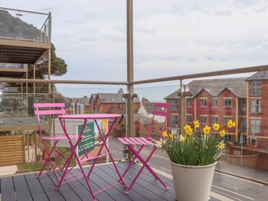 A balcony with pink metal table and chairs and a white pot of yellow flowers overlooking brick buildings at Rolles Rainbow in Exmouth