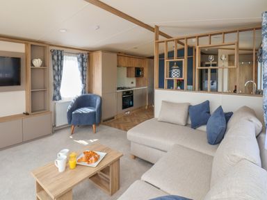 A living room with a beige sofa blue armchair wooden coffee table with croissants and drinks and an open kitchen at Aspen Lodge in Whitby