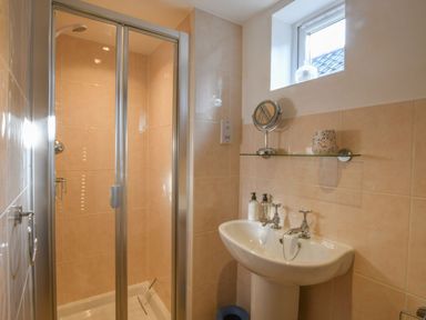 A bathroom with a shower enclosure a sink with two taps and a glass shelf under a window at Caines Cottage in Westleton