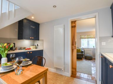 A kitchen with blue cabinets a wooden table set with bowls wine glasses and flowers next to a doorway leading to a living room at Caines Cottage in Westleton