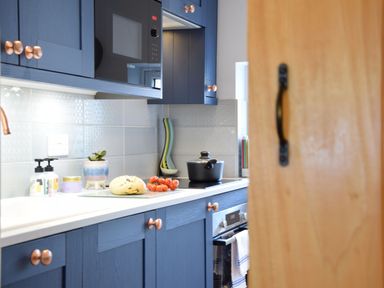 A kitchen with blue cabinets copper handles a microwave a pot on the stove bread and tomatoes on the countertop at Caines Cottage in Westleton