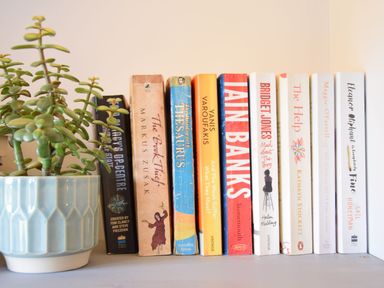 A row of books next to a green plant in a blue pot on a shelf at Caines Cottage in Westleton