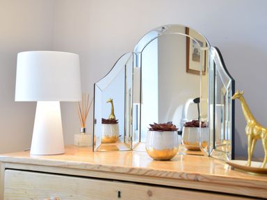 A wooden dresser with a white lamp a glass diffuser a three-panel mirror a small plant pot and a golden giraffe figurine at Caines Cottage in Westleton