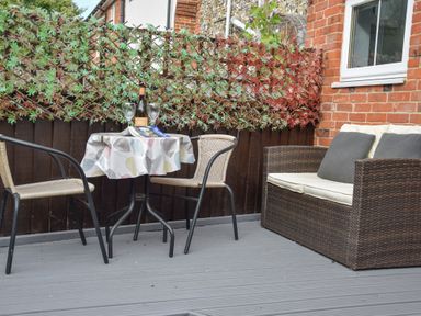 An outdoor patio with two chairs and a table with a cloth a bottle and two glasses and a wicker loveseat with cushions at Caines Cottage in Westleton