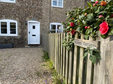 A stone cottage with white door and windows next to a wooden fence with flowers at Caines Cottage in Westleton