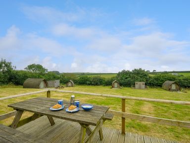 A wooden picnic table with croissants and cups on a deck overlooking a grassy field with small wooden cabins at Hen House @ Penbugle Organic Farm in St Keyne near Duloe