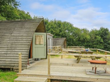 Two wooden pods with a green door on a wooden deck with picnic tables and a fire pit surrounded by trees at Hen House @ Penbugle Organic Farm in St Keyne near Duloe
