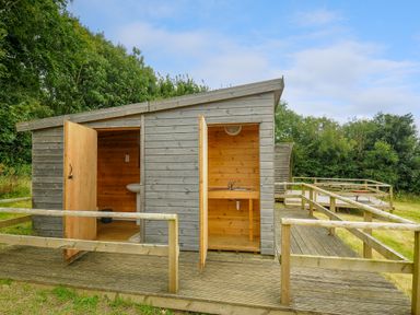 A wooden outdoor structure with open doors revealing a sink and a washbasin surrounded by trees and grass at Pig Pen @ Penbugle Organic Farm in St Keyne near Duloe