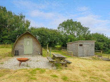 A wooden hut with a door and a green front next to a wooden shed and a picnic table with benches on grass at Pig Pen at Penbugle Organic Farm in St Keyne near Duloe