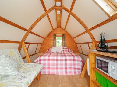 A bedroom with a bed, small window, sofa with cushions, microwave, and kitchen utensils in a wooden cabin at Badger Set @ Penbugle Organic Farm St Keyne near Duloe