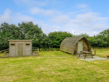 A wooden shed and a curved wooden cabin with picnic table on grass with trees in the background at Badger Set @ Penbugle Organic Farm in St Keyne near Duloe