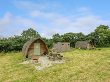 Two wooden pods with picnic tables and a storage shed on a grassy area with trees in the background at Rabbit Warren Penbugle Organic Farm St Keyne near Duloe