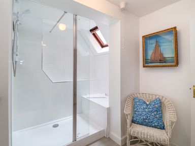 A shower area with glass doors and a wicker chair with a blue pillow next to a framed sailboat painting at 1 Market Hill in Orford