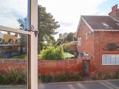 A view through an open window showing a brick house with a tiled roof a brick wall with a wooden gate greenery and trees some houses in the background and a castle in the distance at 1 Market Hill in Orford