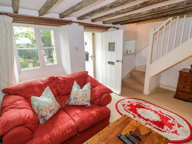 A living room with a red sofa two pillows wooden coffee table with remotes oval rug stairs and a white door at 4 Lansdown Cottages Bourton-On-The-Water