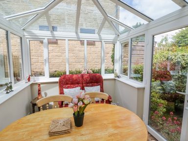 A conservatory with a wooden table with flowers and chairs and a red cushioned bench at 4 Lansdown Cottages in Bourton-On-The-Water