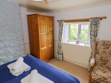 A bedroom with a wooden wardrobe floral armchair and a window with patterned curtains at 4 Lansdown Cottages in Bourton-On-The-Water