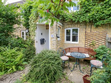 A garden patio with a wooden bench a metal table and two chairs surrounded by plants at 4 Lansdown Cottages in Bourton-On-The-Water