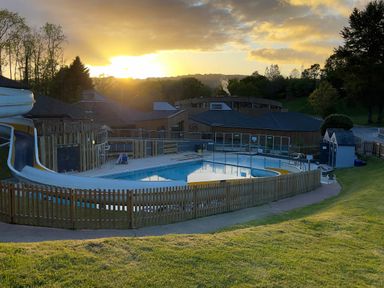 An outdoor swimming pool with a water slide at sunset surrounded by wooden fence and buildings at Lodge Nine Finlake Holiday Park near Chudleigh