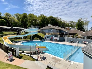 An outdoor swimming pool with a water slide and a lifeguard chair surrounded by buildings and trees at Lodge Nine Finlake Holiday Park near Chudleigh