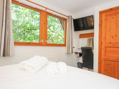 A bedroom with a bed with rolled towels near a window and a wall mounted TV above a desk chair at Lodge Nine in Finlake Holiday Park near Chudleigh