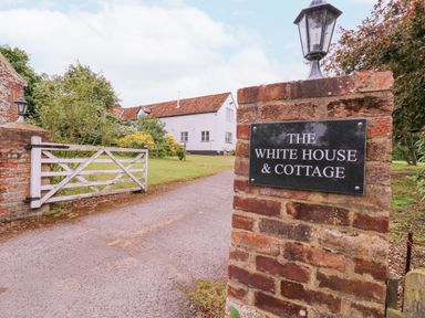 A brick pillar with a sign saying The White House and Cottage next to a white wooden gate and a driveway leading to a white house