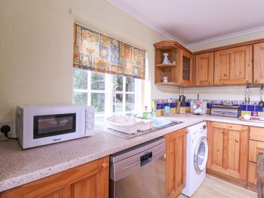 A kitchen with wooden cabinets a microwave dishwasher washing machine and a window with patterned curtains at White House Cottage in Little Snoring near Fakenham