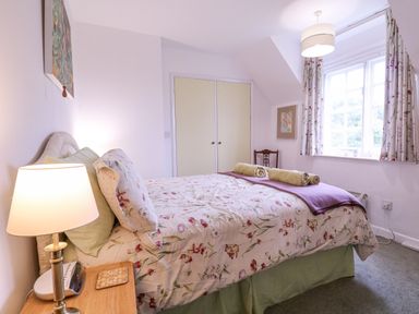 A bedroom with a floral bedspread a bedside table with a lamp and an alarm clock a window with curtains and a chair at White House Cottage in Little Snoring near Fakenham