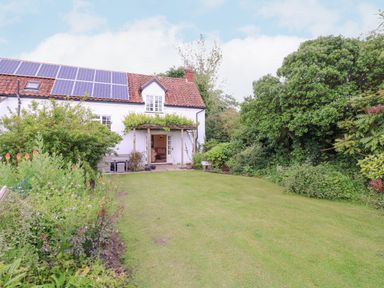 A garden with green lawn and plants in front of a white house with solar panels on the roof at White House Cottage in Little Snoring near Fakenham