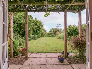 A garden with green lawn and plants viewed through open doors under a wooden pergola at White House Cottage in Little Snoring near Fakenham