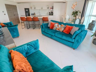 An open living room and kitchen with teal sofas orange cushions kitchen island with orange bar stools and a glass dining table at 17 Cliff Edge in Newquay