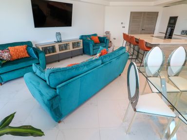 A living room with teal sofas orange cushions a glass dining table with white chairs and an open kitchen with orange bar stools at 17 Cliff Edge in Newquay