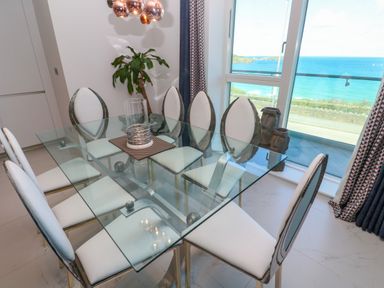 A dining area with a glass table and six white cushioned chairs next to a window overlooking the sea at 17 Cliff Edge in Newquay