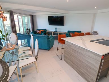 An open plan living area with teal sofas, orange bar stools at a kitchen island, and a glass dining table with white chairs at 17 Cliff Edge in Newquay
