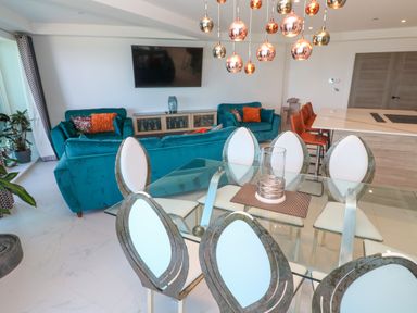 A living room and dining area with teal sofas orange chairs glass dining table and metallic hanging lights at 17 Cliff Edge in Newquay