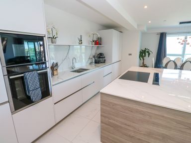 A modern kitchen with white cabinets a built-in oven a sink a kitchen island with a cooktop and a dining area with chairs and a window at 17 Cliff Edge in Newquay