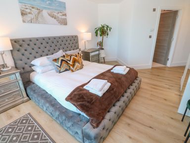 A bedroom with a grey upholstered bed with bedding and pillows beside mirrored nightstands and a plant at 17 Cliff Edge in Newquay