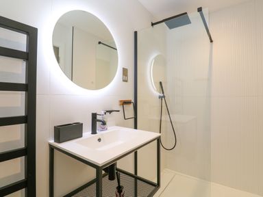 A bathroom with a round illuminated mirror a white sink with black faucet and a glass enclosed shower at 17 Cliff Edge in Newquay