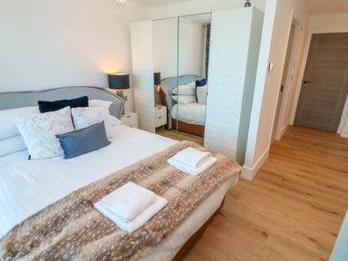 A bedroom with a bed towels pillows a mirrored wardrobe a bedside table with a lamp and wooden flooring at 17 Cliff Edge in Newquay