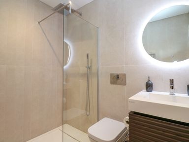 A bathroom with a glass shower enclosure a wall-mounted toilet a vanity with a sink and a round backlit mirror at 17 Cliff Edge in Newquay