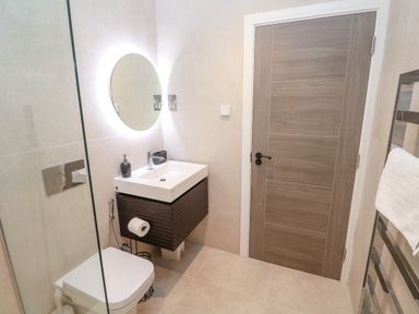 A bathroom with a toilet, a sink with soap dispenser, a round mirror, a wooden door and a towel rack with a white towel at 17 Cliff Edge in Newquay