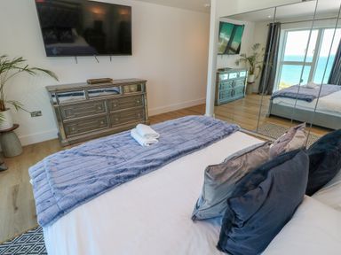 A bedroom with a bed, pillows, a blanket, mirrored wardrobe, chest of drawers, a television mounted on the wall, a plant, and a window with a view of the sea at 17 Cliff Edge in Newquay