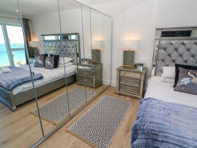 A bedroom with a bed with pillows and a blanket bedside table with a lamp mirrored wardrobe and a patterned rug at 17 Cliff Edge in Newquay