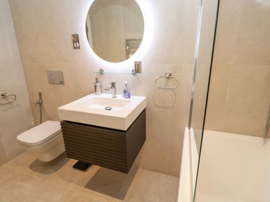 A bathroom with a wall-mounted toilet a sink with a faucet and soap dispensers a round backlit mirror and a bathtub with a glass partition at 17 Cliff Edge in Newquay