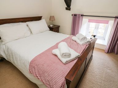 A bedroom with a wooden bed frame white bedding pink blanket and rolled white towels near a window with pink curtains at Rose Hip Cottage in Bellingham