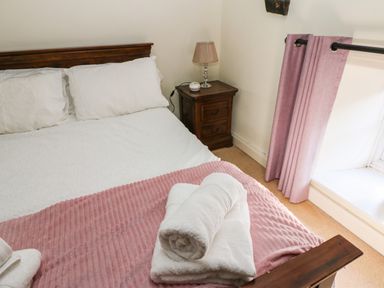 A bedroom with a wooden bed frame white bedding a pink blanket white towels on the bed a wooden bedside table with a lamp and pink curtains at Rose Hip Cottage in Bellingham