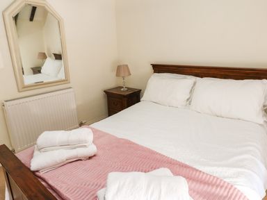 A bedroom with a bed with white linens and a pink blanket a bedside table with a lamp and a wall mirror at Rose Hip Cottage in Bellingham